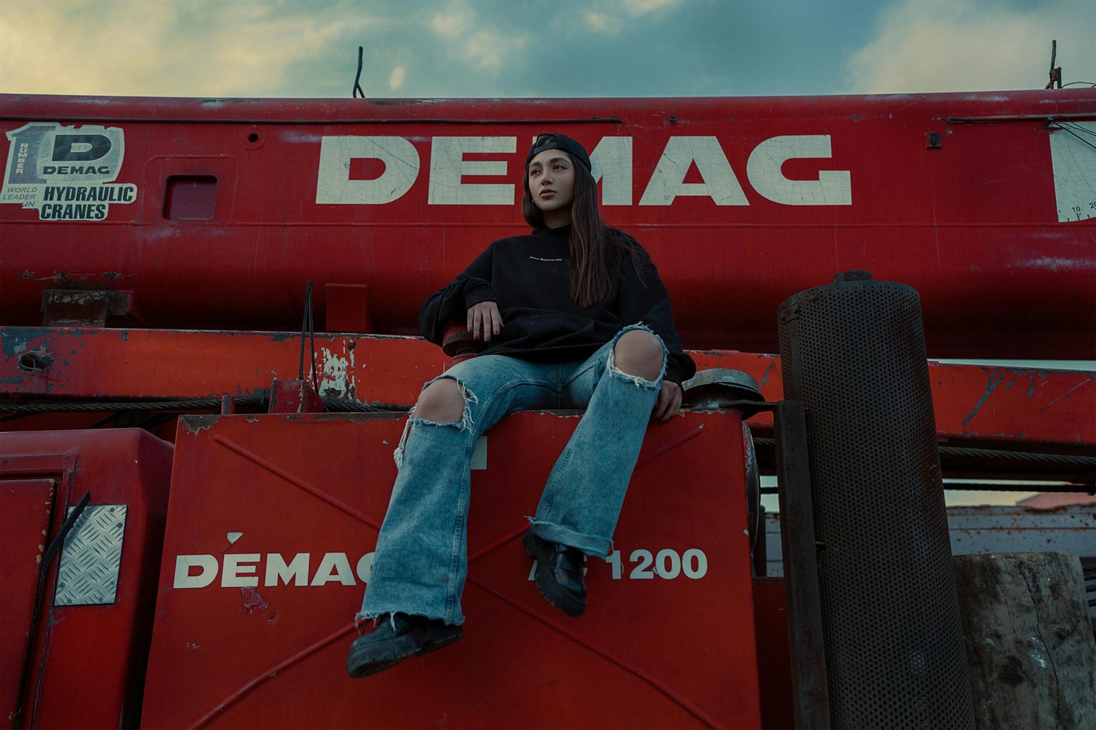 Model poses on industrial equipment, showcasing torn jeans and casual fashion.